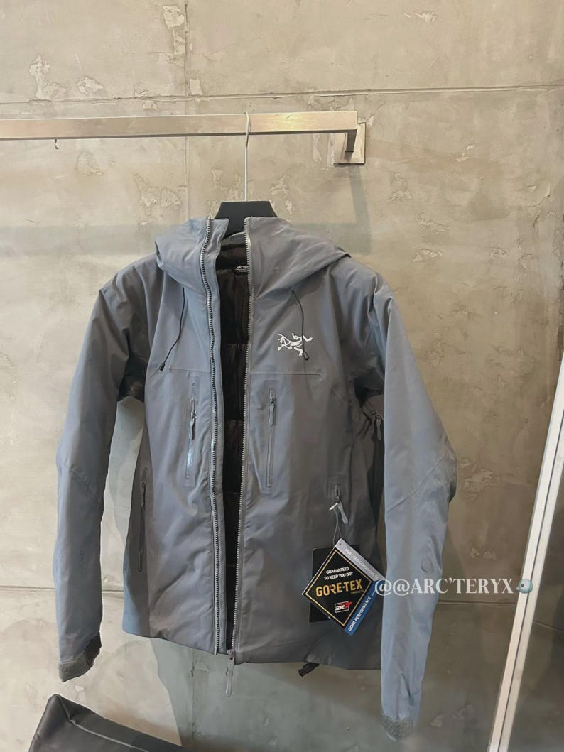 May include: A gray hooded jacket with a zipper closure and multiple pockets. The jacket has a white logo on the chest and a Gore-Tex tag. The jacket is hanging on a metal bar.
