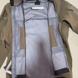 May include: A tan and grey waterproof jacket with a full-length zip and multiple pockets. The jacket has a high collar and a grey interior. The brand name "Arc'teryx" is visible on the collar.