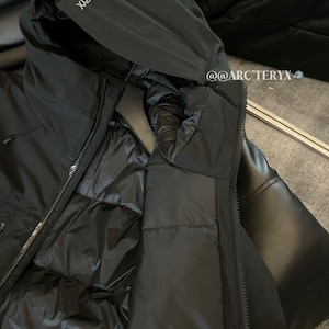 May include: Black hooded jacket with a zipper closure. The jacket features the brand name "Arc'teryx" on the hood. The interior of the jacket is visible, showing a quilted lining. The jacket is displayed on a black surface.