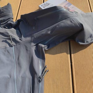 May include: A grey hooded jacket with multiple zipped pockets and drawstrings. The jacket is made of a textured material and is lying on a wooden surface. The text "FULL STOP" is visible on a book in the background.
