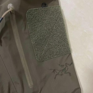 May include: Close-up of olive green outdoor trousers. Features include a zipped pocket, a hook-and-loop patch, and a black logo of a skeleton-like creature. The trousers have a drawstring and a small black loop.