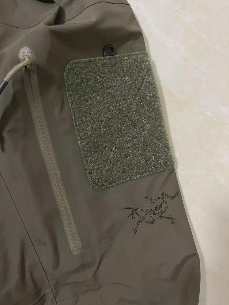 May include: Close-up of olive green outdoor trousers. Features include a zipped pocket, a hook-and-loop patch, and a black logo of a skeleton-like creature. The trousers have a drawstring and a small black loop.