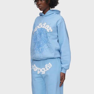 Op de afbeelding: Lichtblauwe hoodie en joggingbroek set. De hoodie heeft een witte spinnenwebafbeelding en het woord "spider" in een gestileerd lettertype. De joggingbroek heeft ook het woord "spider". De outfit is gemaakt van zacht materiaal.