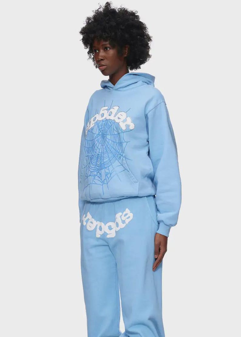 Op de afbeelding: Lichtblauwe hoodie en joggingbroek set. De hoodie heeft een witte spinnenwebafbeelding en het woord "spider" in een gestileerd lettertype. De joggingbroek heeft ook het woord "spider". De outfit is gemaakt van zacht materiaal.