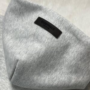 Puede incluir: Primer plano de una sudadera con capucha gris con un peque&ntilde;o parche rectangular de cuero marr&oacute;n oscuro. El parche est&aacute; grabado con la palabra "ESSENTIALS" y la frase "FEAR OF GOD" en letras blancas. La sudadera con capucha est&aacute; hecha de una tela suave y texturizada.