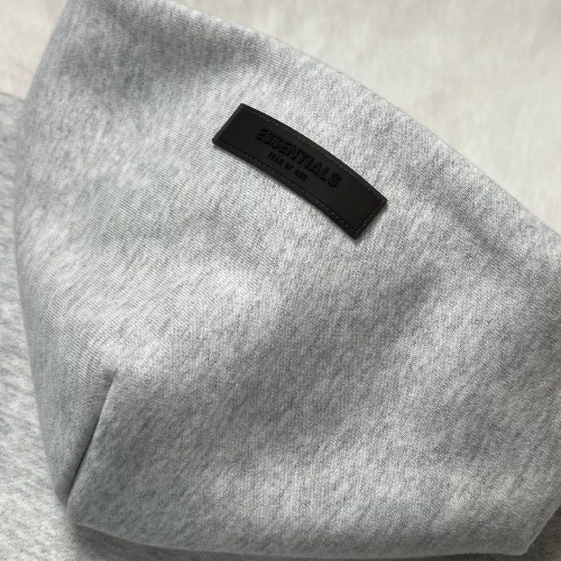 Puede incluir: Primer plano de una sudadera con capucha gris con un peque&ntilde;o parche rectangular de cuero marr&oacute;n oscuro. El parche est&aacute; grabado con la palabra "ESSENTIALS" y la frase "FEAR OF GOD" en letras blancas. La sudadera con capucha est&aacute; hecha de una tela suave y texturizada.