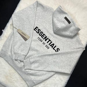 Puede incluir: Una sudadera con capucha gris claro con la palabra "ESSENTIALS" impresa en negro en diagonal en la parte delantera, con "FEAR OF GOD" debajo. Una etiqueta con la palabra "ESSENTIALS" est&aacute; adjunta. La sudadera con capucha est&aacute; hecha de un material suave y c&oacute;modo.