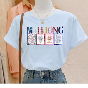 May include: A light blue short-sleeved t-shirt featuring the word MAHJONG in colorful patterned letters above four rectangular stamp-style graphics. The graphics depict mahjong tiles, a blue floral vase, a pink floral vase, and blue floral patterns.