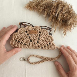 Puede incluir: Un estuche de gafas de ganchillo beige con un borde festoneado, que contiene gafas. El estuche tiene una correa de muñeca con un cierre plateado. Una pequeña etiqueta de madera en el estuche dice "HAND MADE".