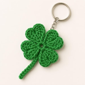 May include: A green four-leaf clover keychain crafted from crocheted yarn. The clover features four rounded, textured petals and a single stem, attached to a circular silver-toned metal split ring by a short chain link connector.
