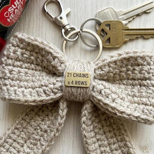 Può includere: Portachiavi a forma di fiocco beige all'uncinetto con chiusura e anelli argentati. Una piccola etichetta di legno recita "21 CHAINS x 4 ROWS". Due chiavi sono visibili sullo sfondo. Accessorio fatto a mano.
