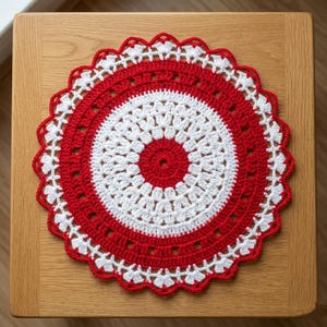 Puede incluir: Un tapete de ganchillo circular hecho a mano en rojo y blanco. El tapete presenta un círculo central rojo, un círculo interior blanco y un anillo exterior rojo, con un borde blanco festoneado. El tapete está colocado sobre una superficie de madera.