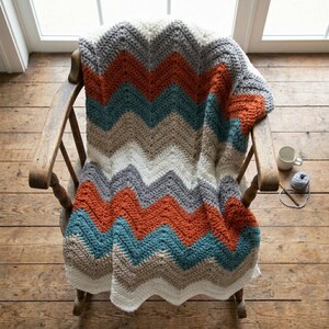 Pode incluir: Uma manta artesanal com padrão chevron em tons de branco, cinza, verde-azulado, laranja e bege está sobre uma cadeira de madeira. O design em zigue-zague adiciona um visual aconchegante e texturizado. Uma caneca e lã com uma agulha de crochê estão por perto.