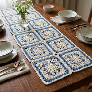 May include: A handmade crochet table runner composed of square motifs. Each square features a cream-colored floral design with a blue border. The runner is displayed on a wooden table, alongside tableware and a vase of flowers.