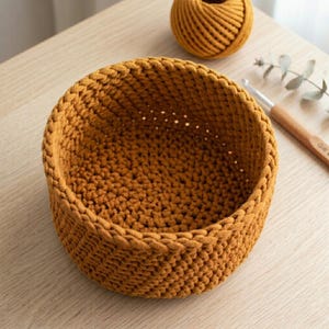 Pode incluir: Uma cesta redonda de crochê feita à mão, na cor amarelo mostarda. A cesta é feita com fio grosso, mostrando um padrão de ponto texturizado. Uma bola de fio correspondente e uma agulha de crochê também são visíveis.