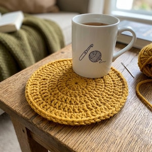 May include: A white mug with a crochet hook and yarn ball design sits on a mustard yellow crocheted coaster. The mug contains a dark liquid. The coaster is on a wooden table.