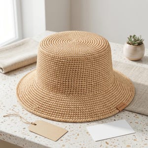 Peut inclure: Un chapeau bob crocheté de couleur beige clair avec un sommet plat et un bord large incliné vers le bas. Le chapeau présente un motif de mailles serrées et uniformes, ainsi qu'une petite étiquette rectangulaire en similicuir marron avec le texte "MAKER".