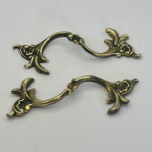 May include: Two antique brass drawer pulls with ornate, curved designs. Each pull features a central handle with decorative floral and leaf accents on the ends. The pulls are made of a shiny, golden-coloured metal.