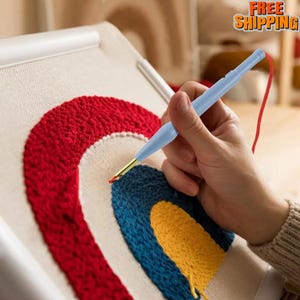 May include: A light blue punch needle tool is positioned over a canvas with a textured rainbow design in red, blue, and yellow. The tool has a gold tip and a red thread. The craft project is in progress.