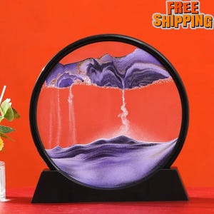 May include: A round sand art piece with a black frame and base. The interior features a vibrant orange background with purple and white sand creating a mountain and waterfall effect. The words "FREE SHIPPING" are in the upper right corner.