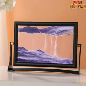 May include: A decorative sand art piece with a black frame and stand. The sand art features purple and white sand, creating a landscape scene against a peach-colored background. The frame has gold-colored accents.