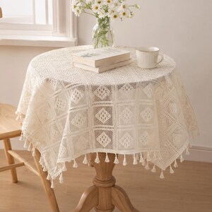 May include: A round, cream-colored crochet tablecloth with a geometric diamond pattern and small tassels along the edge. The tablecloth is draped over a round wooden pedestal table, which also holds a glass vase of white daisies and a white mug.