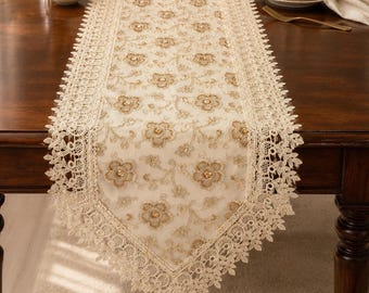 May include: A rectangular table runner featuring an intricate cream-colored lace design with gold floral embroidery and small gold bead accents. The runner has a scalloped lace border and is centered on a dark brown wooden dining table.