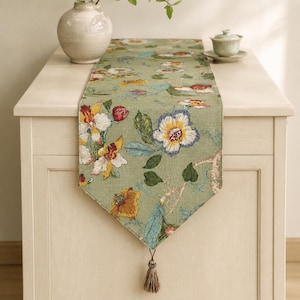 May include: A sage green table runner featuring a multi-colored floral print with white, yellow, and red blossoms and green leaves. The runner has a pointed end finished with a single beige tassel and is draped over a light-colored wooden cabinet.
