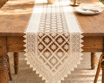 Peut inclure: Un chemin de table en dentelle couleur crème présentant un motif répétitif de losanges avec des fleurs à l'intérieur de chaque forme. Le chemin possède un bord festonné et pointu avec des détails complexes sur une table en bois.