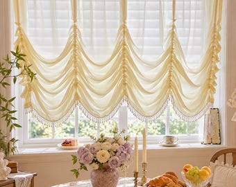 Puede incluir: Una cortina de ventana transparente de color amarillo pálido con paneles de tela fruncidos verticalmente y un borde inferior festoneado adornado con pequeños flecos de cuentas blancas. La cortina cuelga sobre una ventana.