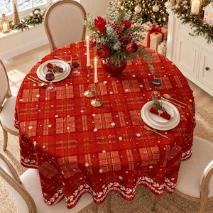 Op de afbeelding: Een rond rood tafelkleed met een ruitpatroon, witte sneeuwvlokaccenten en een decoratieve rand. De tafel is gedekt met witte borden, rode servetten, wijnglazen, goudkleurig bestek en twee hoge kaarsen naast een centraal bloemstuk.