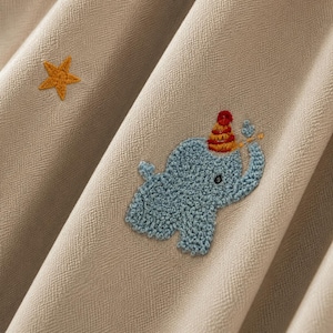 May include: A beige fabric featuring an embroidered light blue elephant wearing a red and yellow striped party hat, accompanied by a small yellow star. The elephant is depicted with a trunk raised, and the fabric has a visible woven texture.