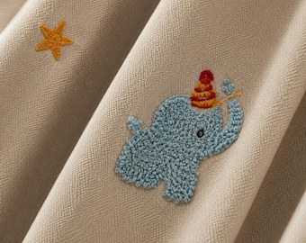 Puede incluir: Una tela de color beige con un elefante bordado en azul claro que lleva un gorro de fiesta a rayas rojas y amarillas, acompañado de una pequeña estrella amarilla. El elefante tiene la trompa levantada y la tela tiene una textura tejida.