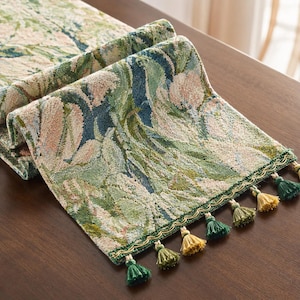 May include: A rectangular table runner featuring a woven tapestry design with floral and leaf patterns in shades of sage green, dark blue, and soft pink. The edges are finished with a decorative gold trim and alternating green and gold tassels.