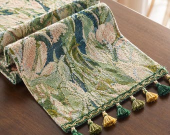May include: A rectangular table runner featuring a woven tapestry design with floral and leaf patterns in shades of sage green, dark blue, and soft pink. The edges are finished with a decorative gold trim and alternating green and gold tassels.