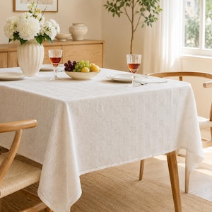Pode incluir: Uma toalha de mesa retangular em tom branco sujo, apresentando um padrão de grelha subtil e repetitivo de pequenos quadrados tecidos. O tecido tem uma aparência texturizada semelhante ao linho e cobre totalmente a superfície da mesa de madeira.