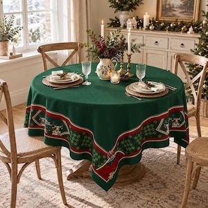 Op de afbeelding: Een rond donkergroen tafelkleed met een decoratieve rand met rode en gouden strepen, een zwart-groen ruitpatroon en rendiermotieven. De tafel is gedekt met witte borden, gevlochten placemats, glazen en een centraal bloemstuk als middelpunt.