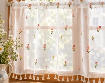 Puede incluir: Una cortina de café blanca y transparente con pequeños motivos florales bordados en rojo, rosa y amarillo dispersos por la tela. La cortina tiene un bolsillo para barra en la parte superior y un dobladillo inferior con borlas blancas.