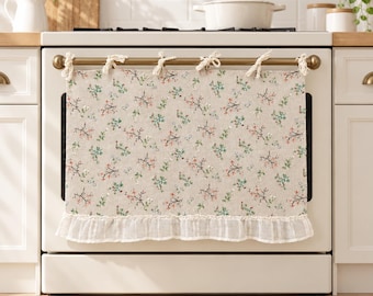 Puede incluir: Un paño de horno beige con un patrón repetitivo de pequeñas ramas florales rojas, verdes y blancas. La tela rectangular se sujeta al asa del horno con cinco lazos de tela anudados y tiene un borde con volantes blancos transparentes.