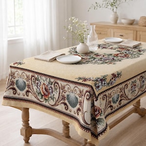 May include: A rectangular beige tablecloth featuring a woven border of floral patterns, roses, and seashell motifs in shades of green, brown, and red. The fabric has a textured appearance with a central floral wreath design and folded napkins.