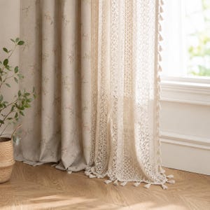 Könnte beinhalten: Ein Vorhangpaar bestehend aus einem beigen Stoffpaneel mit zartem Blumenmuster und einem transparenten weißen Spitzenpaneel mit komplexen geometrischen Details. Das Spitzenpaneel ist mit einer dekorativen Fransenborte versehen.