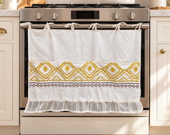 Puede incluir: Una cortina de cocina de tela blanca colgada del tirador del horno con lazos. La cortina presenta una banda horizontal de patrones de diamantes bordados en amarillo, un borde geométrico marrón fino y un dobladillo con volantes transparentes.