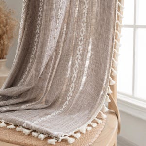 May include: A taupe-colored fabric curtain featuring vertical white embroidered geometric patterns and stripes. The edge of the curtain is finished with a row of small, white decorative tassels that hang along the side and bottom hem.