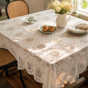 May include: A square, off-white lace tablecloth with a circular pattern covers a wooden table. The table is set with plates, silverware, a teacup, and a small vase of flowers. Wicker chairs surround the table.