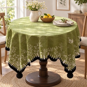 May include: A round green tablecloth featuring a white botanical leaf pattern and a black border with white vertical stripes along the hem. The tablecloth is finished with black pom-pom trim at the corners and drapes over a round wooden table.