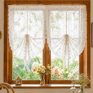 May include: A pair of off-white lace curtains featuring a delicate floral pattern and scalloped edges. The curtains are gathered in the center with vertical ties, creating a fan-like drape that allows light to filter through the intricate fabric.