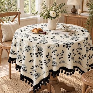 May include: A round tablecloth featuring a cream-coloured base with a black floral rose pattern and a solid black border adorned with hanging black tassels. The tablecloth is draped over a wooden pedestal table, creating a decorative table covering.