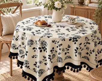 Puede incluir: Un mantel redondo con una base de color crema, un patrón floral de rosas negras y un borde negro sólido adornado con borlas negras colgantes. La tela cubre una mesa redonda de madera, creando una apariencia texturizada y superpuesta.