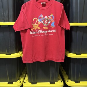 May include: A red short-sleeve t-shirt featuring a graphic of Disney characters Pluto, Donald Duck, Mickey Mouse, and Goofy. The text reads Walt Disney World and Celebrate the Future Hand in Hand. The shirt is displayed on a wooden hanger.