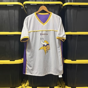 May include: A white V-neck jersey with purple side panels and yellow trim on the collar and sleeves. The front features the word Vikings in purple text above a graphic of a Viking head wearing a horned helmet in purple, yellow, and white colors.